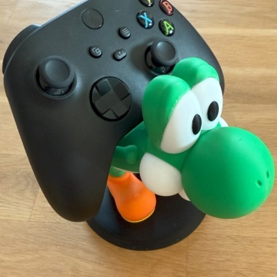 Comando de videojogo preto com botões coloridos e suporte de personagem verde com olhos grandes e sapatos laranja sobre madeira.