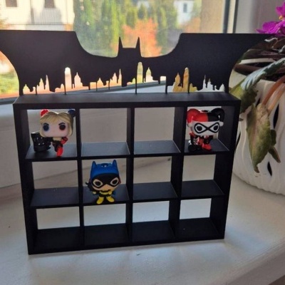 Expositor preto para figuras Funko Pop com símbolo do Batman, com três figuras de personagens em fundo de janela e plantam em vaso branco.