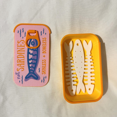 Estojo amarelo aberto com tampa rosa decorada com peixe e texto, com três ossos de peixe brancos dentro
