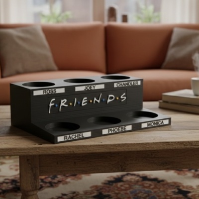 Suporte para copos preto com nomes das personagens da série Friends numa mesa de madeira numa sala com sofá laranja