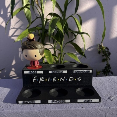 Suporte preto de Funko Pop com nomes dos personagens de Friends e figura Funko Pop de personagem com casaco vermelho.