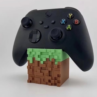 Comando de videojogo preto com botões coloridos apoiado numa base quadrada com padrão pixelado verde e castanho