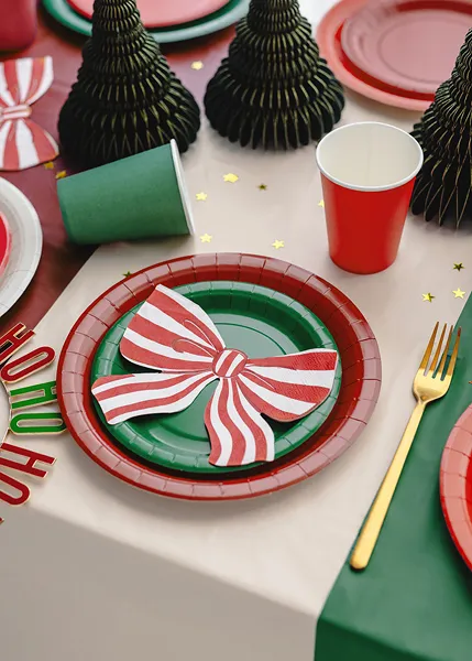 Mesa de Natal com pratos verdes e vermelhos, copos vermelho e verde, laço às riscas brancas e vermelhas, talheres dourados e árvores de papel pretas