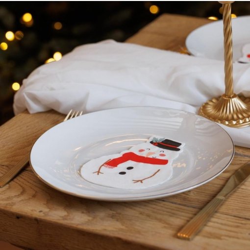 Prato branco com desenho de boneco de neve sobre mesa de madeira com talheres dourados e guardanapo branco.