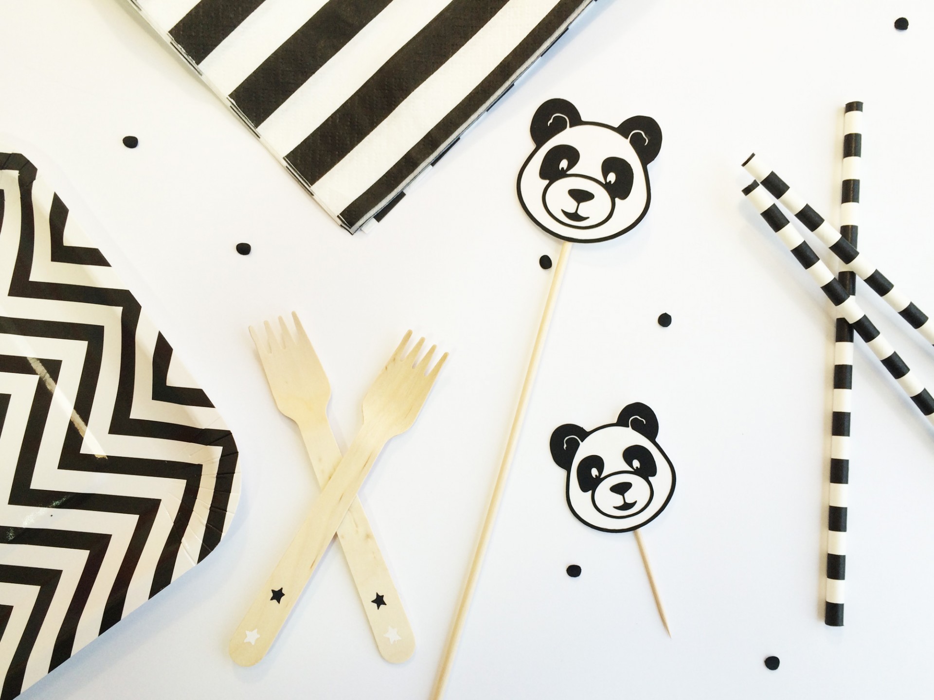 Talheres de madeira, pratos e guardanapos a preto e branco, palhinhas e palitos decorativos de panda numa mesa branca.