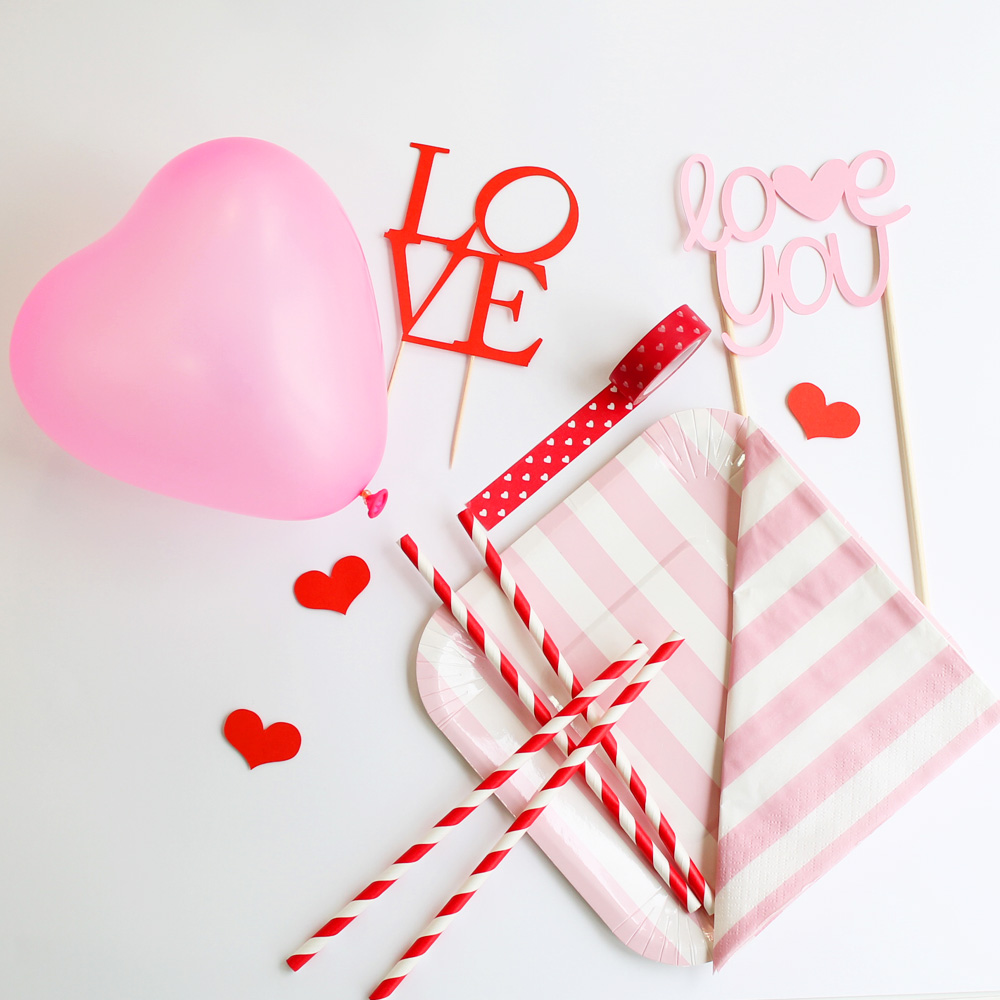 Decoração de festa com balão cor de rosa em forma de coração, pratos, guardanapos riscas rosa e branco, canudos vermelhos com branco e enfeites de madeira com texto LOVE e love you.