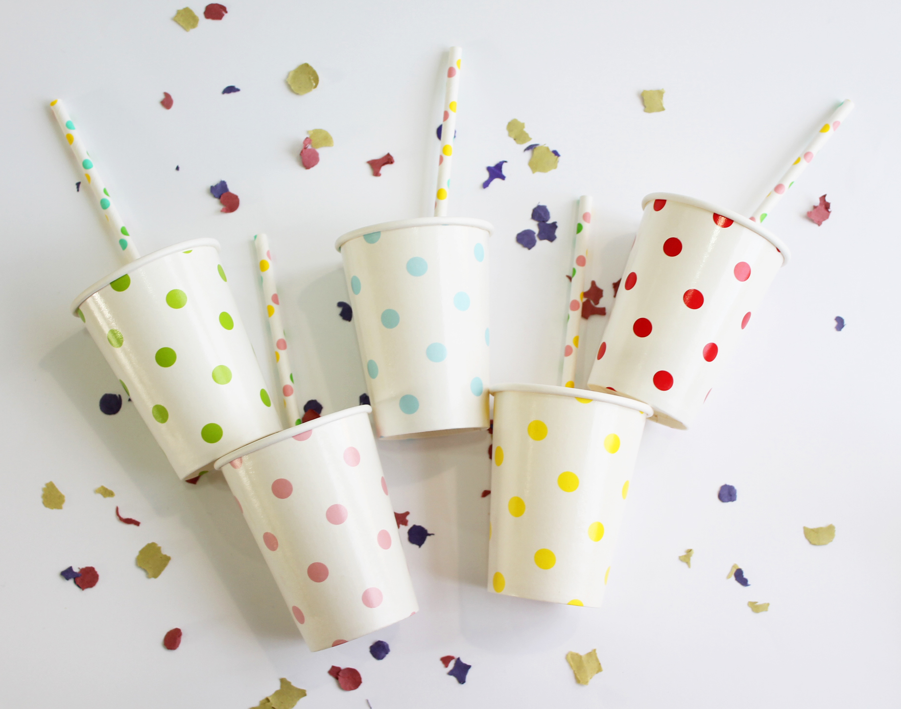 Cinco copos com bolinhas coloridas e canudos, sobre fundo branco com confetes