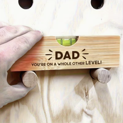 Nível de bolha em madeira clara com gravação para pai, segurado por mão masculina