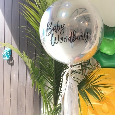 Balão prateado redondo com texto Baby Woodbury e fitas brancas em ambiente com plantas verdes
