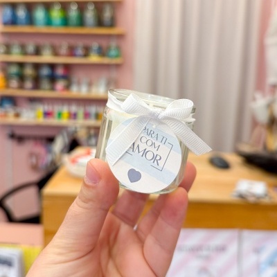 Vela em frasco de vidro com fita e etiqueta, mãos segurando, fundo de prateleiras coloridas e mesa