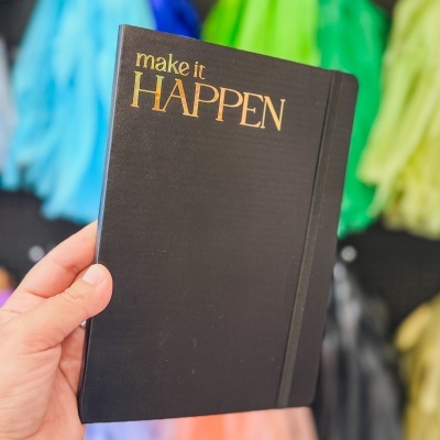 Caderno preto com texto dourado 'make it HAPPEN' na capa.