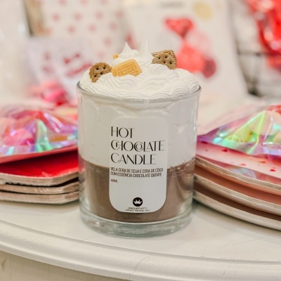 Vela em copo de vidro com decoração que imita chocolate quente com chantilly e biscoitos, sobre mesa branca com pratos coloridos ao fundo