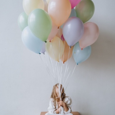 Criança segurando balões coloridos pastel sentada numa caixa contra parede branca