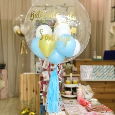 Balão decorativo transparente com balões azuis, brancos e dourados e texto 'Batizado do Santiago' em loja
