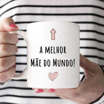 Caneca branca com texto 'A MELHOR MÃE DO MUNDO!' e símbolos, segurada por pessoa com unhas cor de rosa