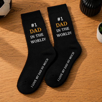 Par de meias pretas escritas #1 DAD IN THE WORLD! e I LOVE MY DAD SO MUCH sobre mesa de madeira com bola de futebol, vaso preto e planta suculenta.