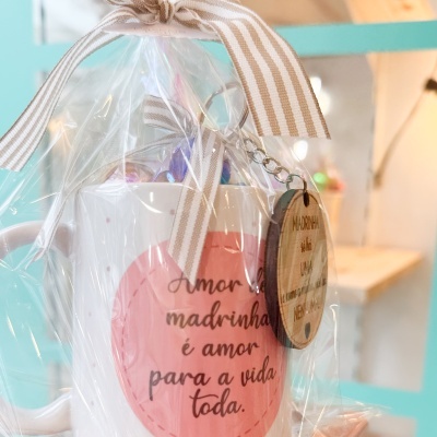 Caneca branca com círculo rosa e texto, embalada em plástico com fita às riscas e pendente de madeira