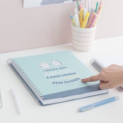 Caderno azul claro com texto em português numa mesa branca com caneca de canetas coloridas e mão apontando