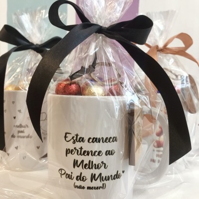 Caneca branca com mensagem em português embalada em celofane com laço preto, exibida em conjunto com outras canecas similares a fundo branco e cores roxas e azul claro.