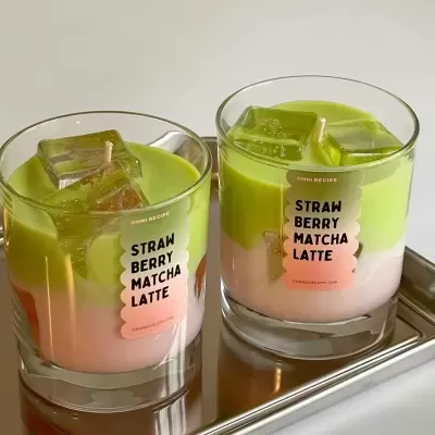 Duas velas em copos de vidro com cera verde e rosa em camadas e cubos transparentes, com etiquetas com texto, numa bandeja metálica.