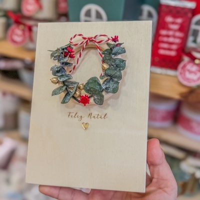 Placa decorativa de Natal em madeira clara com coroa de folhas e flores e texto Feliz Natal