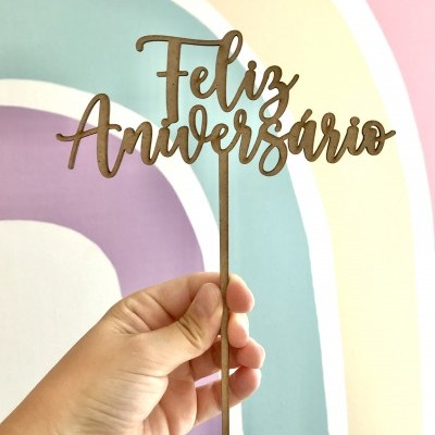 Topo de bolo em madeira com o texto Feliz Aniversário a ser segurado por uma mão em fundo colorido.