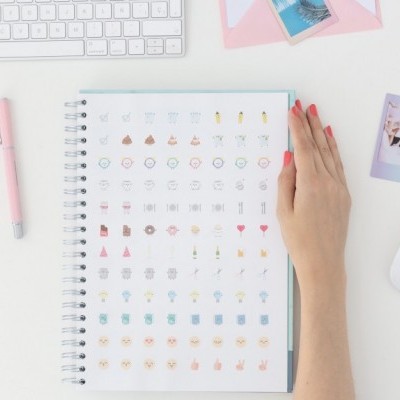 Mão com unhas vermelhas sobre caderno com várias ilustrações pequenas em mesa branca com teclado e caneta