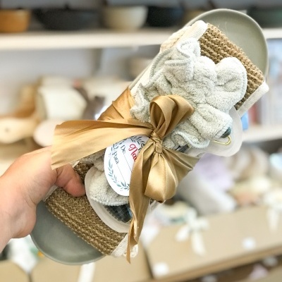 Chinelos cinzentos com tiras castanhas de ráfia e toalha cinzenta decorativa, com laço castanho e etiqueta redonda, em fundo de loja desfocado.