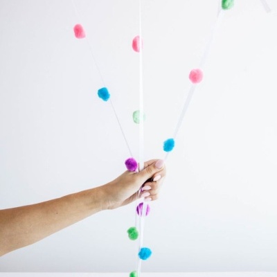 Mão segurando balões cor-de-rosa com fitas decoradas com pompons coloridos.