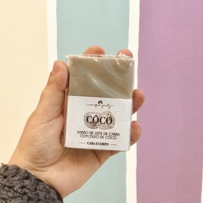 Sabonete coco em embalagem branca segurado por mão com casaco cinzento, fundo colorido