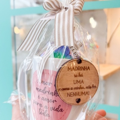 Copo branco com coração rosa e texto para madrinha, embrulhado com fita e medalha de madeira