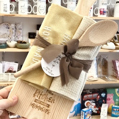 Conjunto de cozinha em madeira e tecido com texto gravado e toalhas enroladas, exposto numa loja com canecas ao fundo.