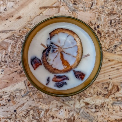 Vela decorativa branca em recipiente cerâmico com frutas secas em cima e madeira reciclada ao fundo