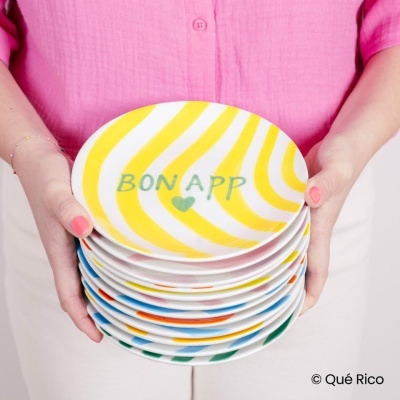 Conjunto de pratos coloridos com riscas amarelas e texto BON APP, segurados por pessoa com camisa rosa
