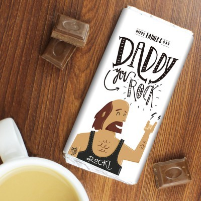 Barra de chocolate com embalagem temática 'Happy Father's Day' e ilustração de homem rockeiro sobre uma mesa de madeira com pedaços de chocolate e chávena branca.