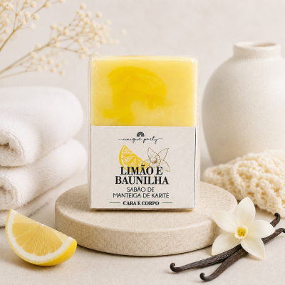 Sabão sólido amarelo e branco com embalagem branca e texto, colocado sobre suporte redondo com flores, baunilha, limão e toalhas brancas.