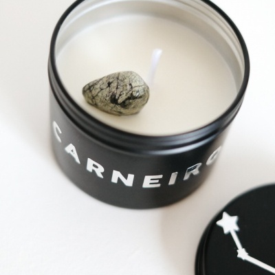 Vela branca em recipiente preto com texto 'CARNEIRO' e tampa preta com desenho de constelação.