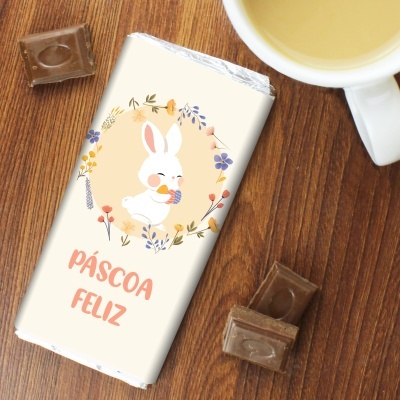 Barra de chocolate embrulhada com design de coelho e texto Páscoa Feliz junto de chávena e pedaços de chocolate