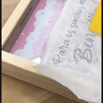 Caixa de madeira com tampa de vidro contendo cartão rosa com nuvens e papel com texto