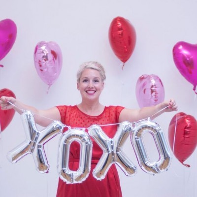 Mulher com vestido vermelho segura balões prateados XOXO com balões corações ao fundo