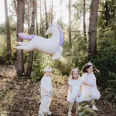 Três crianças vestidas com roupas de festa brancas num bosque com balão gigante de unicórnio
