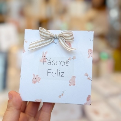 Caixinha de papel branca com padrão de coelhos e texto Páscoa Feliz com laço no topo