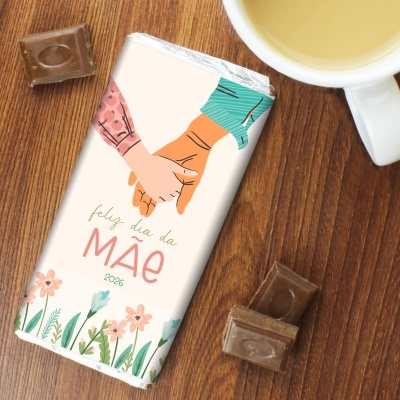 Barra de chocolate com embalagem de dia da mãe, flores e mãos dadas, ao lado de pedaços de chocolate e uma chávena com chá.
