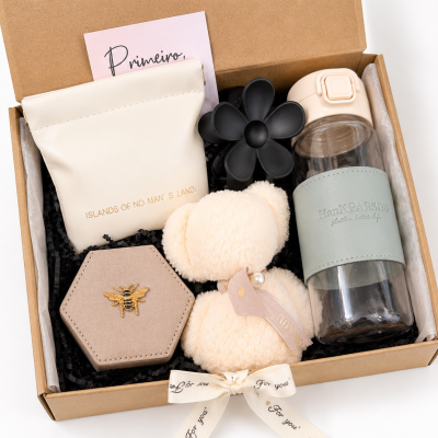 Conjunto de oferta com bolsa branca, caixa bege hexagonal com abelha, garrafa de vidro com capa azul, flor preta e ursinho peluche creme em caixa de cartão