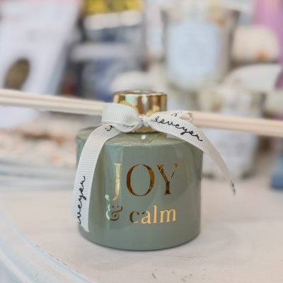 Frasco verde com tampa dourada e inscrições douradas JOY & calm com fita branca com texto preto