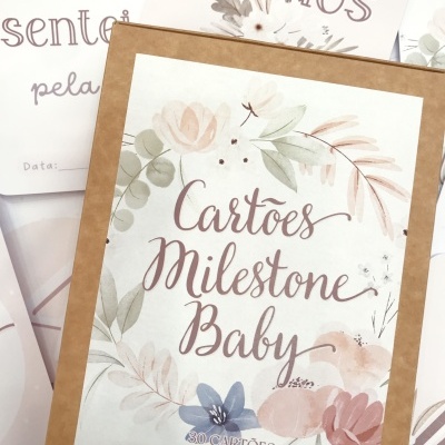 Caixa de cartão decorada com flores e texto Cartões Milestone Baby com cartões ao fundo em tom pastel.