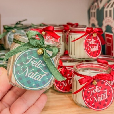 Vela em pote de vidro com etiqueta 'Feliz Natal' e fita verde ou vermelha com sino.