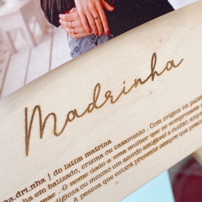 Placa com gravação 'Madrinha' em madeira clara com pessoas abraçadas ao fundo