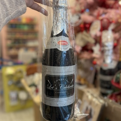 Garrafa negra com rótulo cinzento e texto de celebração embalada em plástico transparente, segurada pela mão com fundo desfocado colorido.