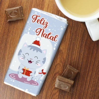 Barra de chocolate com embalagem azul clara e gato de Natal, junto a pedaços de chocolate e chávena numa mesa de madeira.
